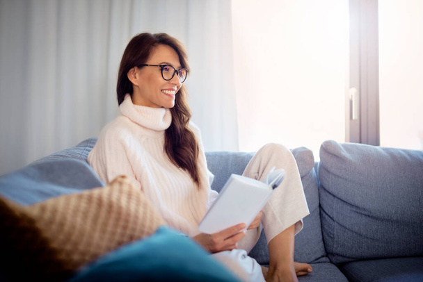 Snímek s úsměvem žena na sobě ležérní oblečení při relaxaci na pohovce doma a čtení knihy. - Fotografie, Obrázek