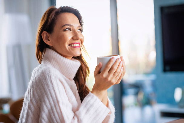Shot of happy attractive woman wearing casual clothes and holding a cup of tea in her hand while relaxing at home.  - Foto, imagen
