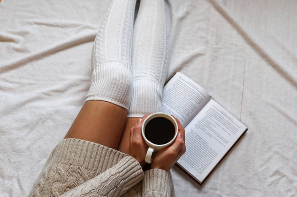 Kobieta trzyma filiżankę kawy na nogach w wełnianych skarpetkach zimowych w przytulnym łóżku z książką w domu. Zbliżenie, wybiórcze skupienie - Zdjęcie, obraz