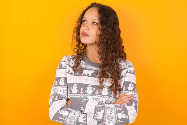 Charming thoughtful  woman wearing Christmas sweater agaist yellow studio background stands with arms folded concentrated somewhere with pensive expression thinks what to do - Photo, Image