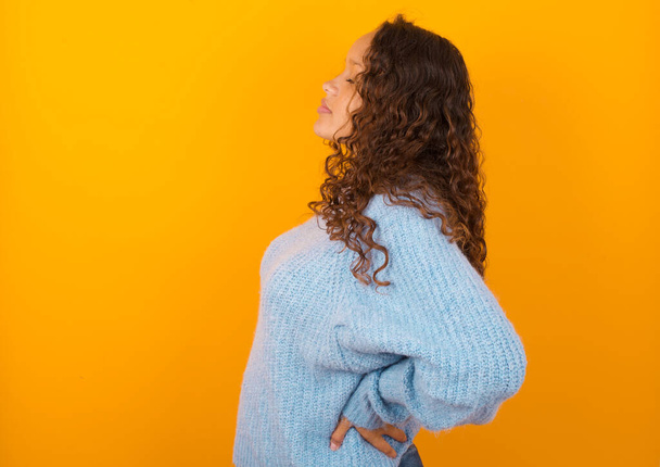 Die niedliche lockige Frau in blauem Pullover über gelbem Studiohintergrund bekam Rückenschmerzen. Studioaufnahme - Foto, Bild