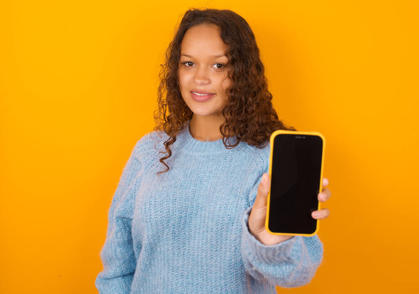 Okouzlující rozkošné kudrnaté žena na sobě modrý svetr přes žluté studio pozadí drží moderní zařízení, ukazuje černý displej smartphone - Fotografie, Obrázek