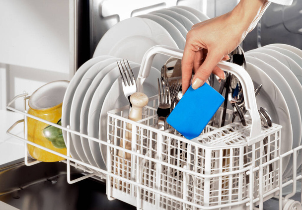 рука женщины в посудомоечной машине, полной посуды и столовых приборов - Фото, изображение