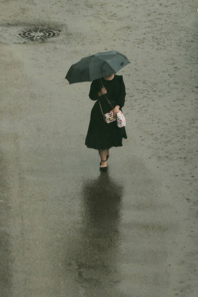 A woman in a black dress and with a black umbrella walks in the rain. A woman's reflection and raindrops on the wet asphalt. A gray autumn day. Top view from the front - Foto, immagini