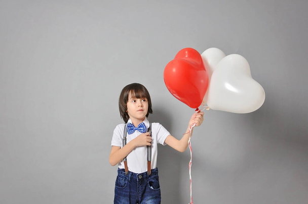 a caucasian European boy is thinking, holding heart-shaped balloons and looking at them. The concept of childhood and happiness. Studio shoot - Zdjęcie, obraz