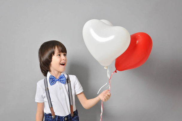 adorable niño caucásico sonriendo, sosteniendo globos en forma de corazón y mirándolos. El concepto de infancia y felicidad. - Foto, Imagen
