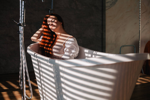 A young, slender, red-haired girl in a black bathroom robe stands in the sunlight through blinds - Foto, afbeelding