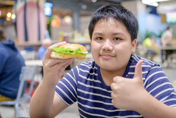 Obézní chlapec si vychutnat vepřový hamburger v jídelně v nákupním centru. koncepce nezdravého jídla a nezdravého jídla - Fotografie, Obrázek