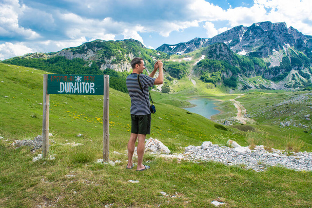 Ein Touristenmann macht ein Foto, während er neben einem Schild mit der Aufschrift Durmitor auf dem Hintergrund wunderschöner Berggipfel steht. Durmitor National Park. Höchster Straßenpass Montenegros im Norden Montenegros. - Foto, Bild