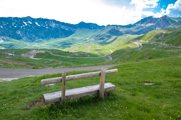 モンテネグロで最も高い道路のパスであるデュルモーター国立公園の山々や道路の背景にある緑の丘の上に木製のベンチ。美しい夏の曇りの風景. - 写真・画像