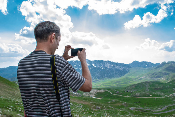 男の観光客は美しい夏の風景の写真を撮る。デュルモーター国立公園の山の峰、緑の丘と曇った青空。モンテネグロで最も高い道路パス. - 写真・画像