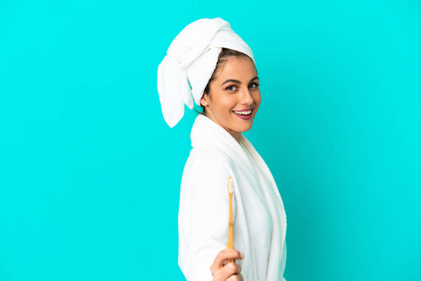 Young woman in a bathrobe brushing teeth over isolated blue background with happy expression - 写真・画像
