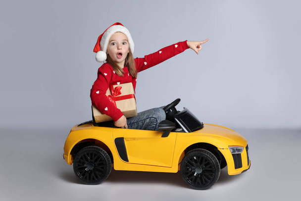 Cute little girl in Santa hat with Christmas gift box driving children's electric toy car on white background - Foto, afbeelding