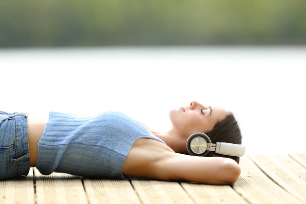 Side view portrait of a relaxed woman wearing headphones listening to music lying on a pier - Foto, Imagem
