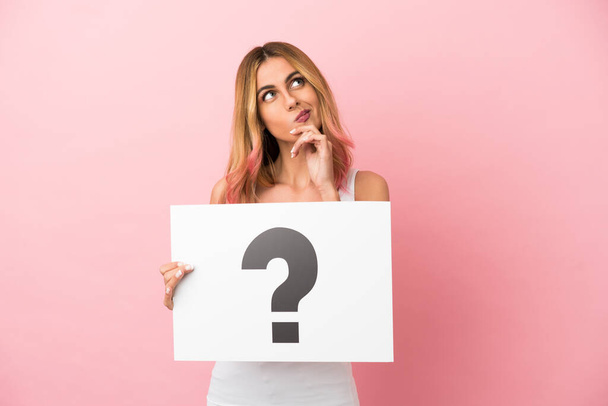 Young woman over isolated pink background holding a placard with question mark symbol and thinking - Фото, изображение
