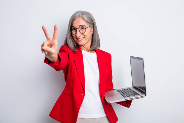 senior pretty woman smiling and looking happy, gesturing victory or peace. business and laptop concept - Foto, Imagen