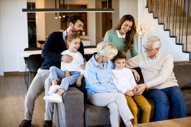 Modern evdeki kanepeye oturup televizyon izleyen çok nesilli bir aile. - Fotoğraf, Görsel