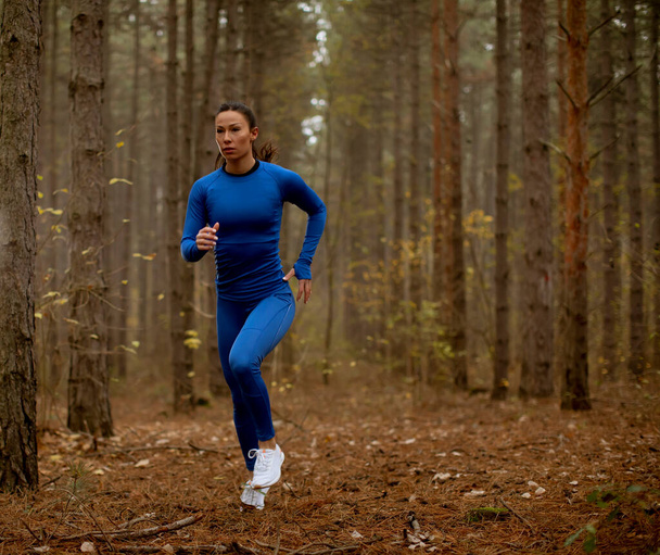 Fiatal nő kék melegítőben fut a kamera felé az erdei ösvényen ősszel - Fotó, kép