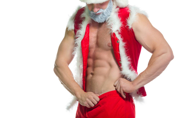 Weihnachten. Porträt eines muskulösen sexy starken Athleten im Weihnachtsmannkostüm mit grauem Bart, der seinen Bauch isoliert auf weißem Hintergrund zeigt - Foto, Bild
