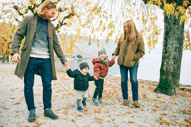 Retrato al aire libre de la joven familia feliz de cuatro, madre y padre jugando con los niños en el parque de otoño junto al lago, clima frío - Foto, imagen