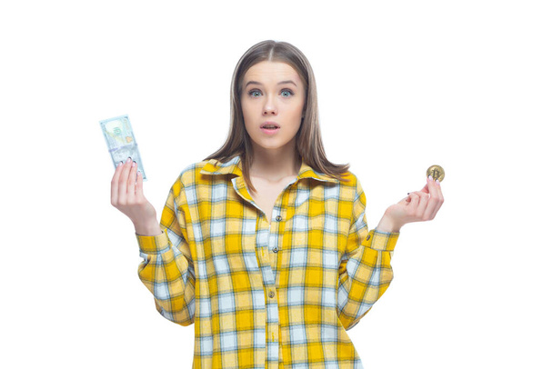 Shot of an astonished young female holding a golden bitcoin in one hand and dollar banknote in the other hand. Doubts where to invest, the choice of currency - Φωτογραφία, εικόνα