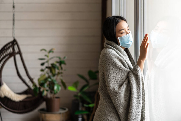 Nemocná Asiatka zůstává doma v karanténě z covid-19, nosí masku, dívá se z okna, touží jít ven - Fotografie, Obrázek