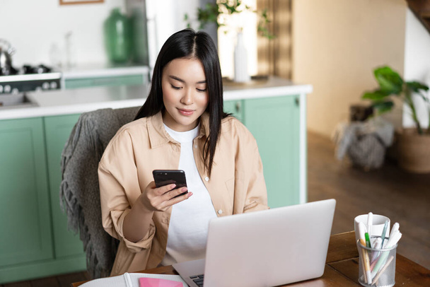 Mujer asiática joven mirando su teléfono, sentada en casa con el ordenador portátil, trabajando o estudiando a distancia. Concepto de educación independiente y a distancia, cursos en línea - Foto, Imagen