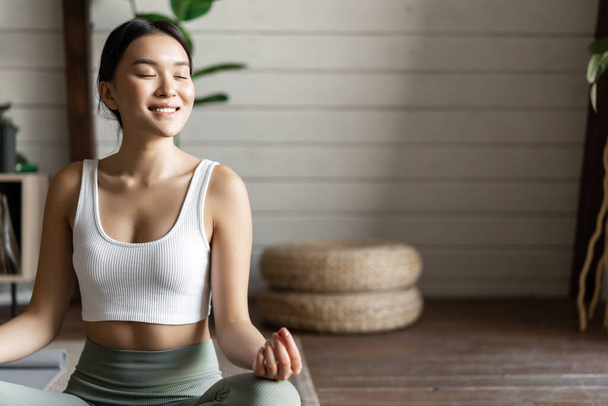 Mindfulness en meditatie concept. Jonge aziatische vrouw doet workout thuis, yoga meditatie in de woonkamer op vloermat, steeds gefocust - Foto, afbeelding
