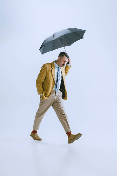 Full-length portrait of young man dressed in 50s, 60s style walking with umbrella isolated on white background. - Zdjęcie, obraz