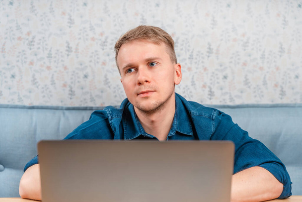 Een jongeman zit aan een tafel en kijkt uit onder een laptop. Werken op een laptop op afstand van huis of van een café. Technologieën, telewerken en sociale netwerken - Foto, afbeelding