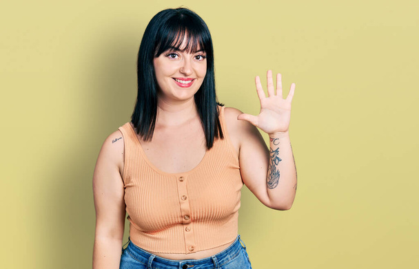 Junge hispanische Plus-Size-Frau trägt lässige Kleidung und zeigt mit Finger Nummer fünf nach oben, während sie selbstbewusst und glücklich lächelt.  - Foto, Bild