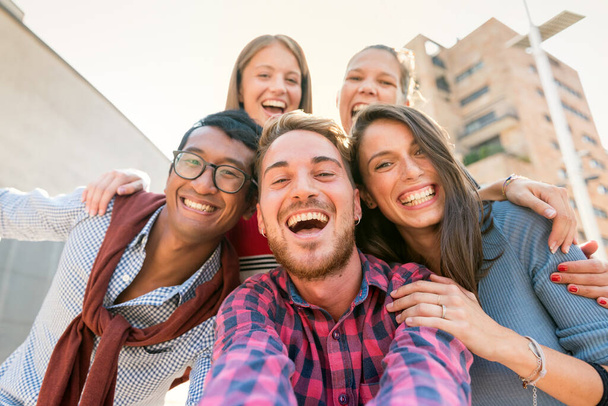 Низкоугольный портрет веселой межрасовой группы из пяти друзей, улыбающихся во время фотографирования на открытом воздухе в городе - Фото, изображение