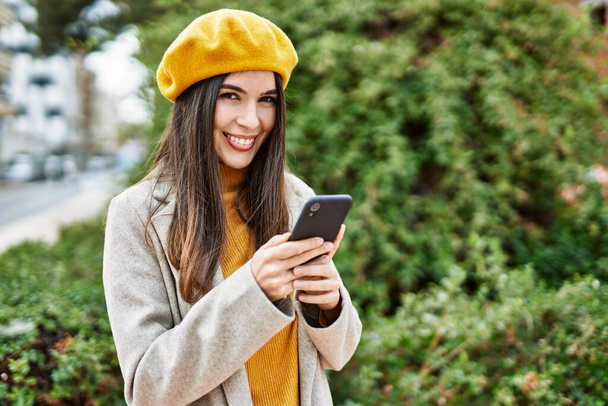 Νεαρή ισπανόφωνη κοπέλα χαμογελά ευτυχισμένη χρησιμοποιώντας smartphone στην πόλη. - Φωτογραφία, εικόνα