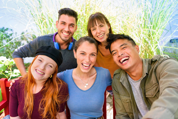 Grupo de alegres amigos masculinos y femeninos multiétnicos en ropa casual sonriendo y mirando a la cámara mientras se toma selfie en el jardín verde en un día soleado - Foto, Imagen