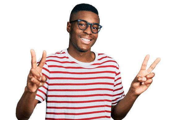 Jeune homme afro-américain portant un t-shirt décontracté et des lunettes souriant en regardant la caméra montrant des doigts faire signe de victoire. numéro deux.  - Photo, image