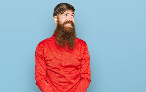 Рыжий мужчина с длинной бородой в повседневной одежде смотрит в сторону с улыбкой на лице, естественным выражением лица. смеясь уверенно.  - Фото, изображение