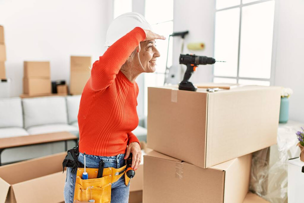 Middle age grey-haired woman wearing hardhat standing at new home very happy and smiling looking far away with hand over head. searching concept.  - 写真・画像