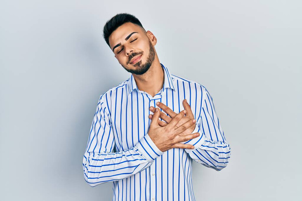 Jeune homme hispanique barbu portant une chemise rayée décontractée souriant avec les mains sur la poitrine avec les yeux fermés et geste reconnaissant sur le visage. concept de santé.  - Photo, image