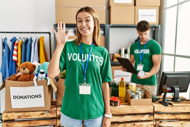 Joven chica rubia vistiendo camiseta voluntaria en el puesto de donación renunciando a decir hola feliz y sonriente, gesto de bienvenida amistoso  - Foto, imagen