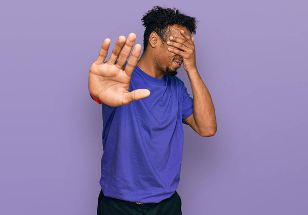 Young african american man with beard wearing casual purple t shirt covering eyes with hands and doing stop gesture with sad and fear expression. embarrassed and negative concept.  - Foto, Bild