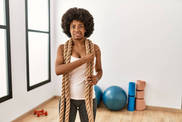 Молода афроамериканська жінка з волоссям афро під час тренувань у спортзалі з бойовими мотузками, що вказують убік стурбовані і знервовані вказівним пальцем, стурбовані і здивовані виразом  - Фото, зображення