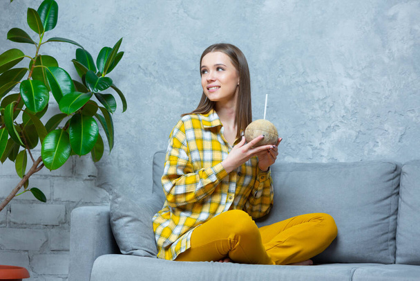 Front view of a smiling beautiful girl sitting cross legged on a sofa at home holding a coconut in both hands looking aside. Concept of healthy lifestyle, nutrition, detox, vegetarianism - Foto, Imagen