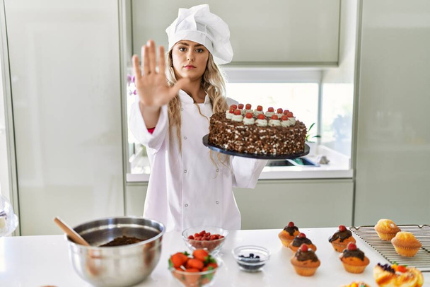 Молодой кавказский кондитер-повар женщина готовит выпечку и торт на кухне с открытой рукой делает знак стоп с серьезным и уверенным выражением лица, защитный жест  - Фото, изображение