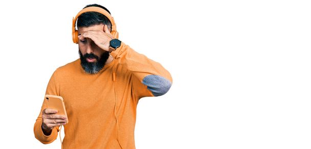 Hispanic man with beard using smartphone wearing headphones stressed and frustrated with hand on head, surprised and angry face  - Фото, зображення