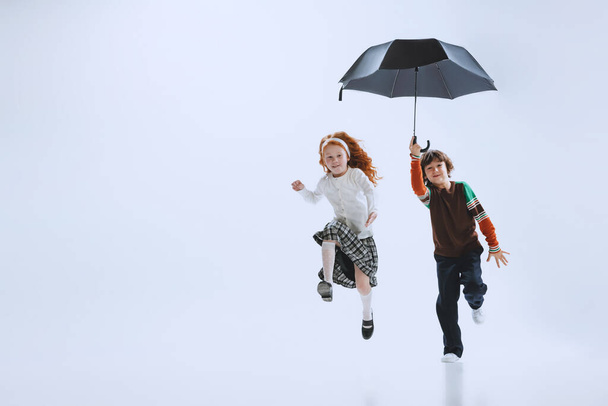 Two happy kids, boy and girl running with umbrella isolated on white studio background. Childhood, dreams, imagination, education - Фото, зображення