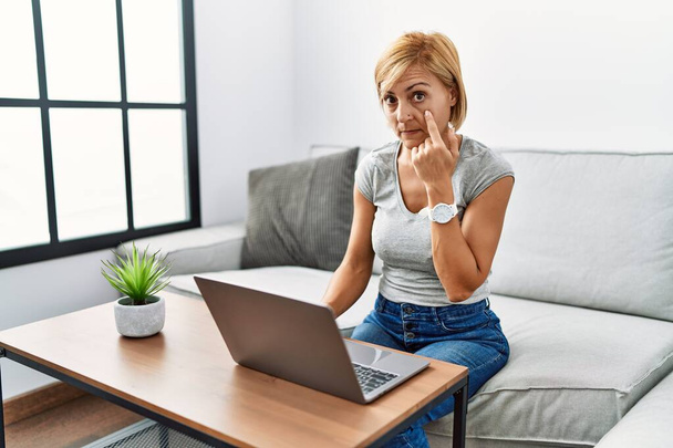 Middelbare leeftijd blonde vrouw met behulp van laptop thuis wijzen naar het oog kijken naar je gebaar, verdachte uitdrukking  - Foto, afbeelding