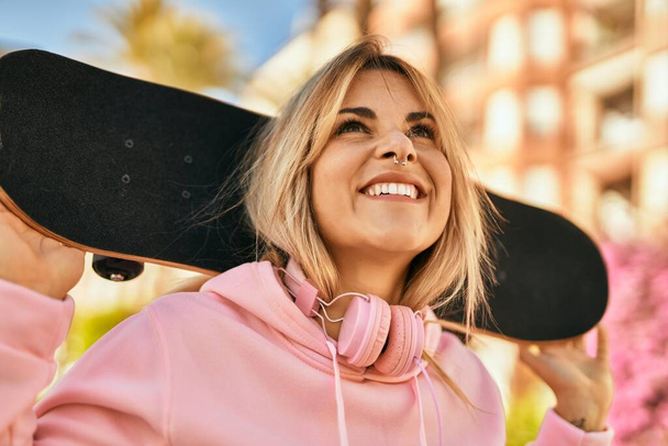 Young blonde skater girl smiling happy holding skate at the city. - Photo, Image
