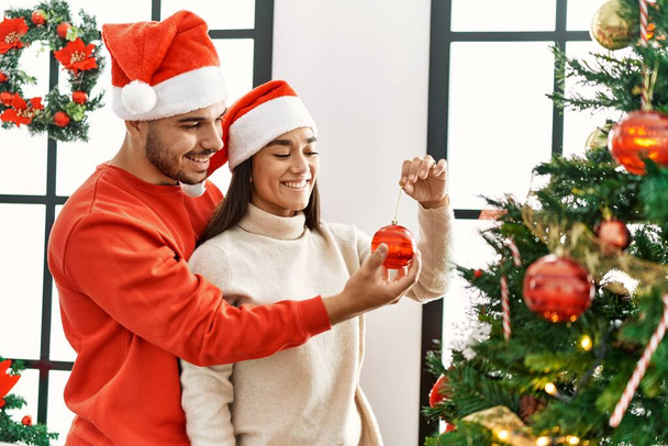 Νεαρό ζευγάρι ισπανόφωνων χαμογελά ευτυχισμένη διακόσμηση χριστουγεννιάτικο δέντρο στο σπίτι. - Φωτογραφία, εικόνα