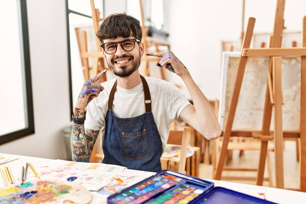 Sanat stüdyosunda sakallı, neşeli gülümseyen, parmakları ve ağzıyla işaret eden İspanyol bir adam. diş sağlığı kavramı.  - Fotoğraf, Görsel