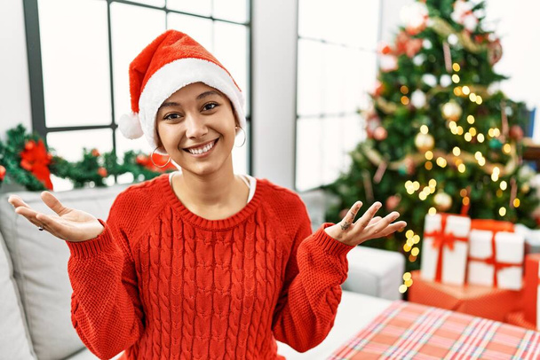 Young hispanic woman with short hair wearing christmas hat sitting on the sofa smiling cheerful with open arms as friendly welcome, positive and confident greetings  - Foto, Imagen